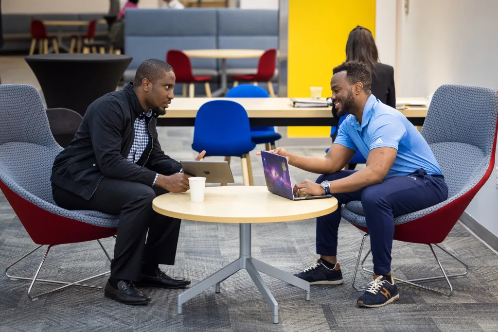 Two students talking at table and using a laptop.